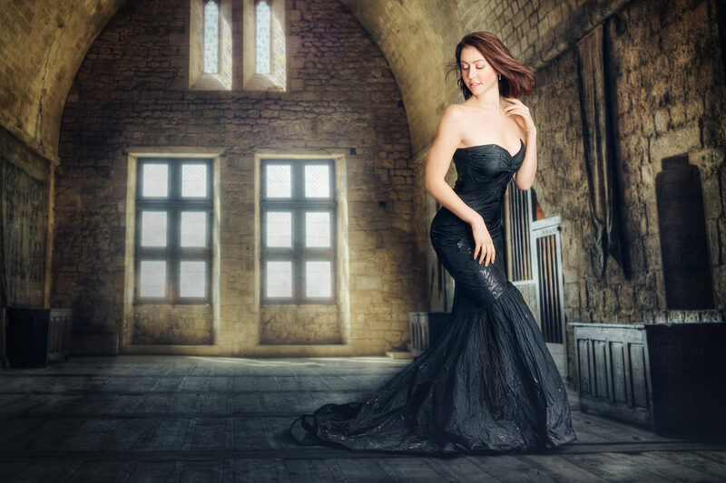 Fine art portrait of an elegant woman in a flowing black gown inside a grand stone hall with tall windows